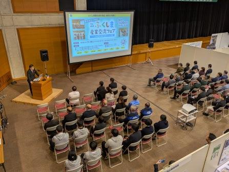 前方中央に資料が映し出された大きなスクリーンが設置され、左側に設置された壇上に立つ市長の話を会場に設置された椅子に座った参加者の方々が聞いている写真