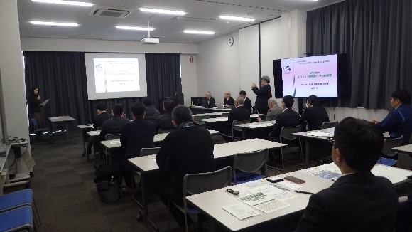 室内前方と右側に資料が映し出されたスクリーンが設置され、スクリーンの左側に女性が立ち、中央の席から立ち上がった男性が話をしている様子の写真