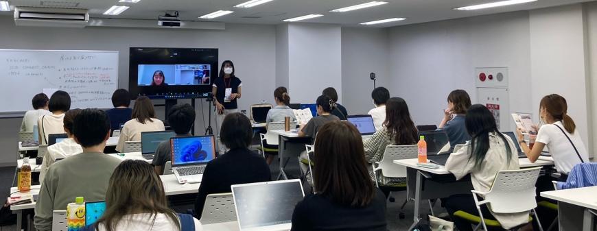 前方に設置された大型モニターに女性講師の顔が表示され、モニターの右側に女性が立ち話をしているのを2名ずつ座った参加者たちが熱心に聞いている講座の様子の写真