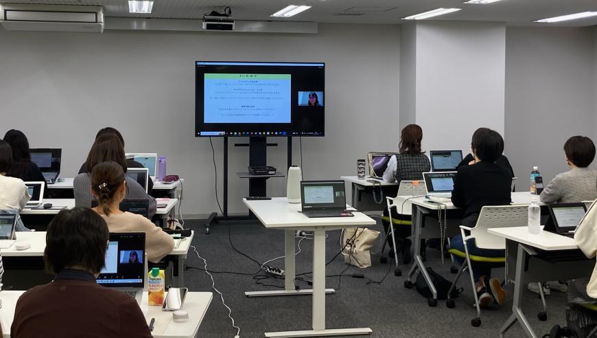 前方に資料が映し出されたモニターが設置され、2名ずつ長机の席に座った参加者たちが受講している様子の写真