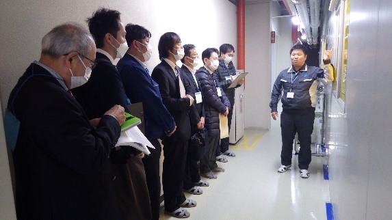 通路のような場所で7名の参加者が縦一列に並び、右側に立っている男性スタッフの説明を聞いている写真
