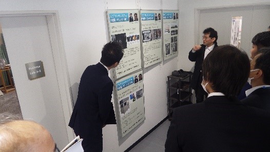 通路の白い壁に4つの大きな商品紹介のポスターが掲示され、右側に立つ男性の話を参加者が聞いている写真