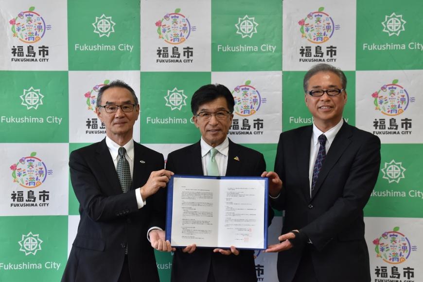 福島市のバックパネルの前に立って協定書をもってカメラに向かって見せている河原田代表取締役社長、木幡市長、乾支店長の写真