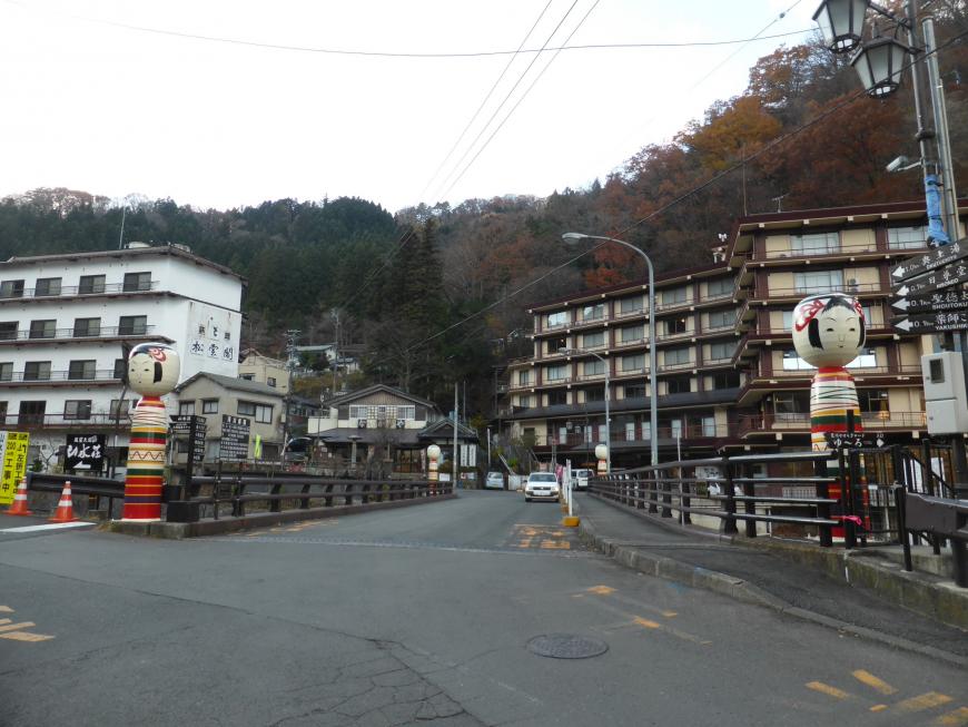 奥に温泉街の旅館やホテルが立ち並び、荒川橋の両脇にこけしのオブジェが立っている写真