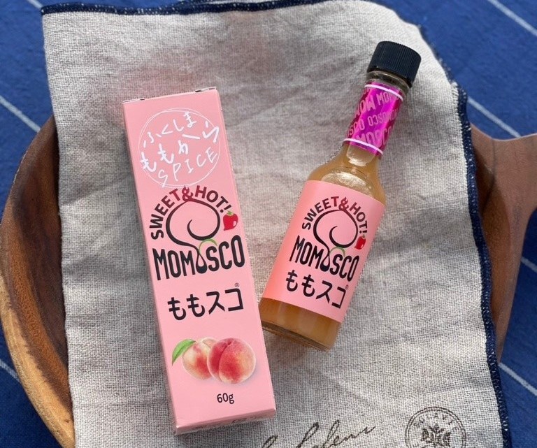木皿に敷かれた布の上に、ピンク色のももスコの箱と、箱と同じデザインの商品シールが貼られた瓶に入ったももスコが並んで置かれている写真