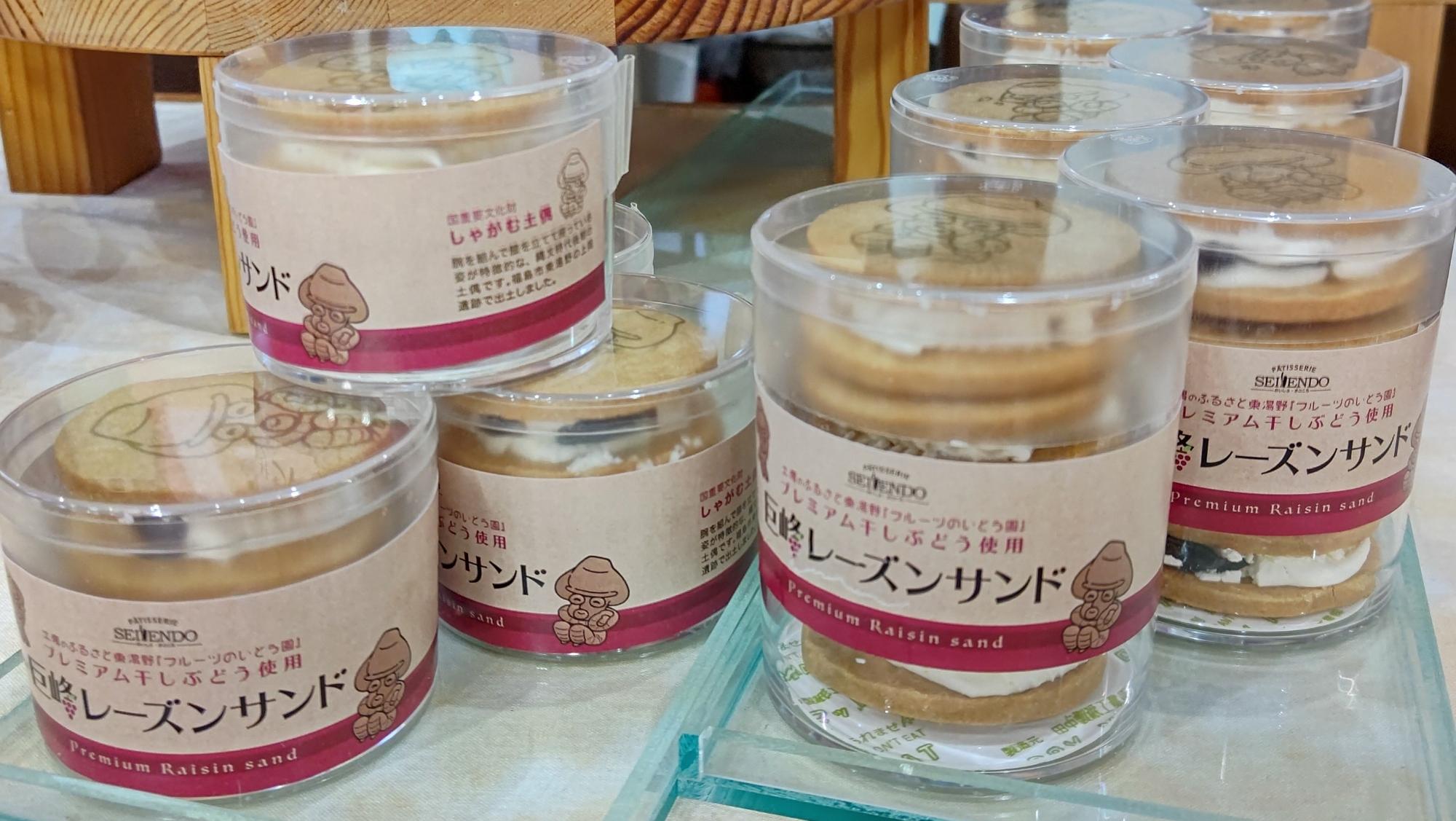 円柱型の透明のケースに「しゃがむ土偶のレーズンサンド」と書かれたラベルが貼られ、クッキーが入った商品が並んで置かれている写真