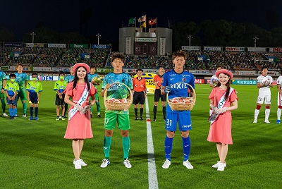 スタジアムの芝にひかれた白いラインの左右に桃が入ったかごを持った選手2名が立ち、その横にピンク色の帽子とワンピースを着た女性が一人ずつ立っている写真