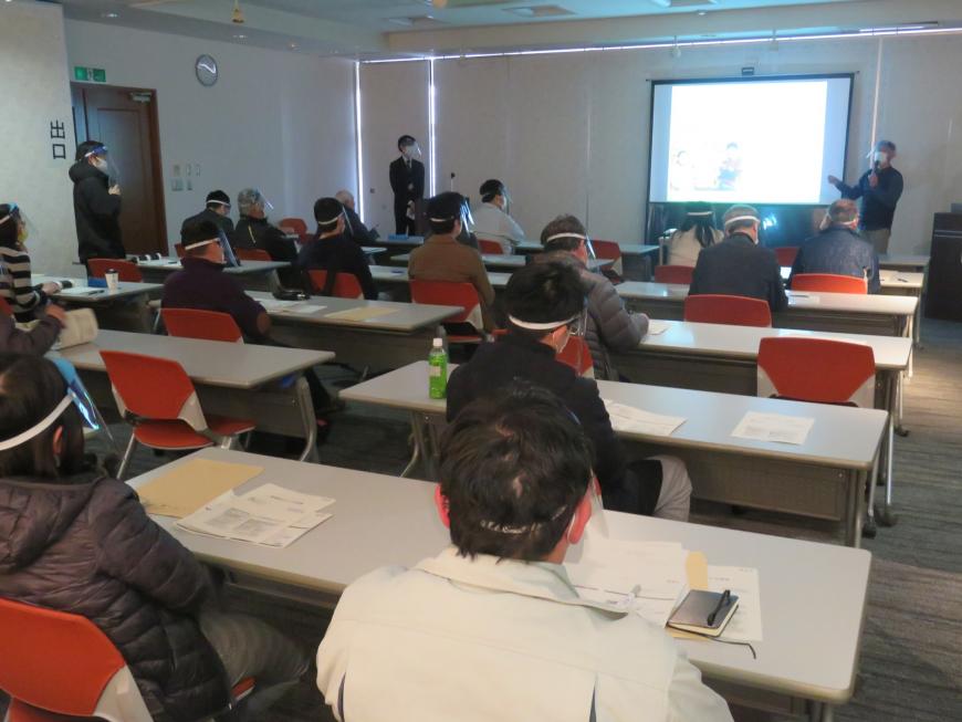 長机の席に参加者たちが座り、前方の資料が映し出されたスクリーン横に立った男性がマイクで話をしている様子の写真