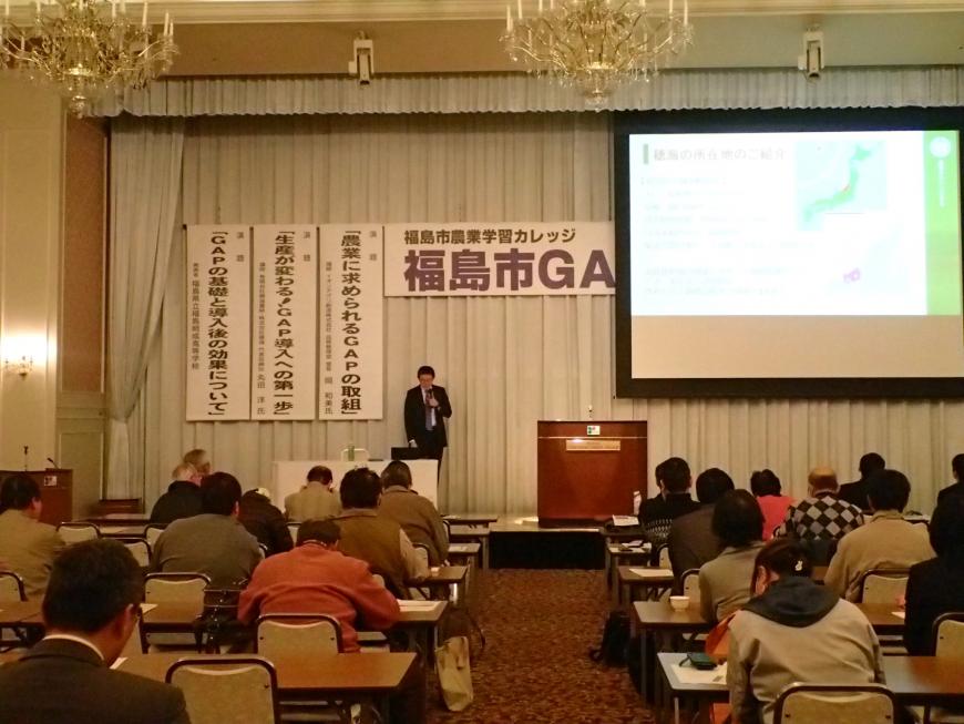 たくさんの参加者たちが席に座る中、舞台の上に設置されたスクリーンにスライドが映し出され、マイクを持った男性が話をしている様子の写真