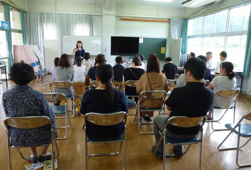 教室に並んだ椅子に受講者たちが座り、前方のホワイトボードの前に立つ女性の方を見ている写真