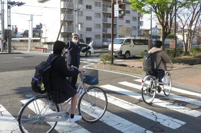紺色のユニフォームを着た交通教育専門員の男性が、横断歩道の横に立ち、学生が乗った2台の自転車の通行の安全を確保している様子の写真