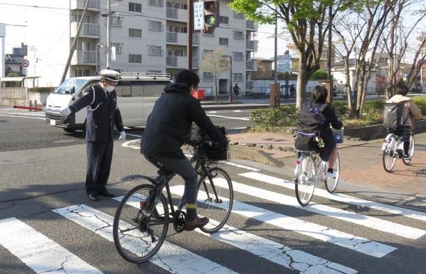 紺色のユニフォームを着た交通教育専門員の男性が横断歩道の横に立ち、通行する3台の自転車の安全を確保している様子の写真