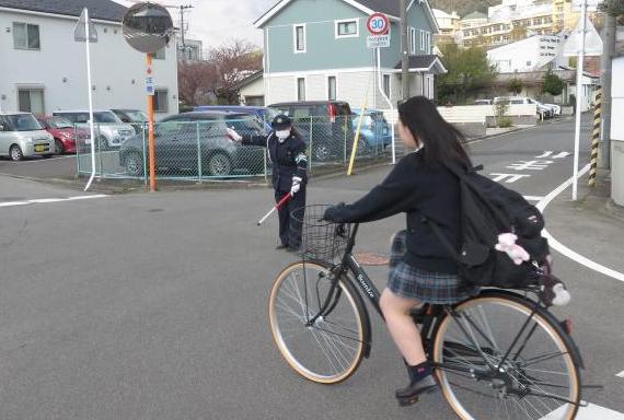 紺色のユニフォームを着た交通教育専門員の女性が、交差点の中に立ち、右側から交差点に進入してくる自転車に乗った女子高校生のために安全を確保している様子の写真