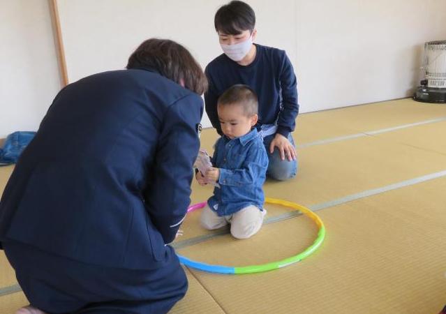 座敷に置かれたフラフープの輪の中に座った小さな男の子と外側に座った男性が、紺色のユニフォームを着た交通教育専門員の女性の話を聞いている様子の写真