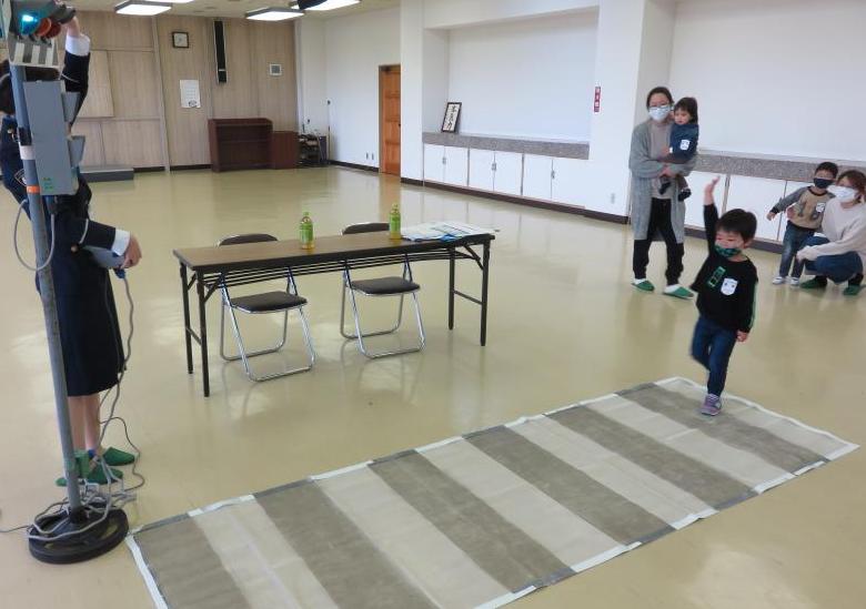 室内に置かれた長机の前に横断歩道のシートが敷かれ、交通教育専門員の女性に倣って右手を挙げた小さな男の子が横断歩道を渡っている様子を2組の親子が見ている写真