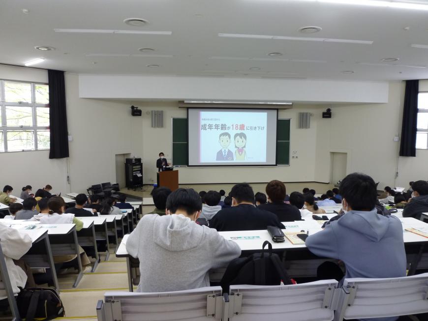 階段状になった席に大勢の学生が座り、スクリーンが設置された黒板前に講師が立ち講演をおこなっている出前講座の様子の写真