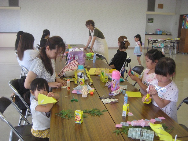 黄色の紙や糊などがたくさん置かれた長机の席に座った親子が七夕飾りの工作をしている様子の写真