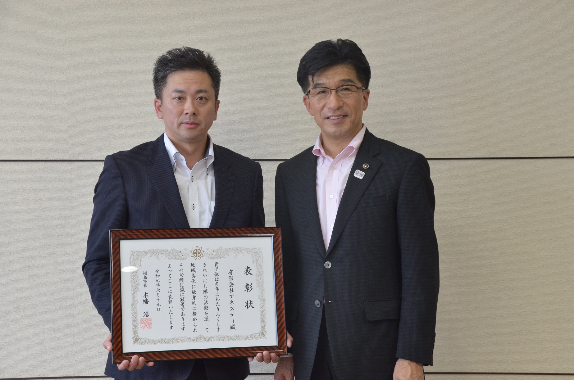 スーツを着た男性が額縁に入った表彰状を持ち、市長と記念撮影している写真