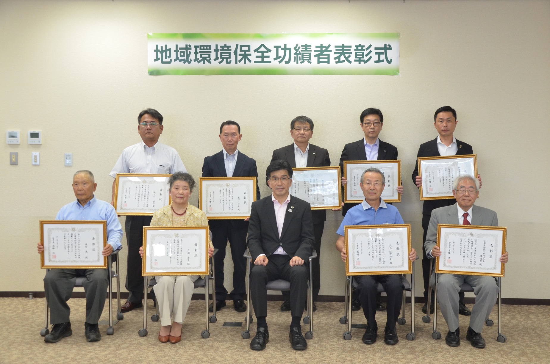 「地域環境保全功績者表彰式」と書かれた垂れ幕の下で前列に市長の両隣に2人ずつ椅子に座り、後列には5名立って並び、表彰者全員が額縁に入った表彰状を手に持ち記念撮影している写真