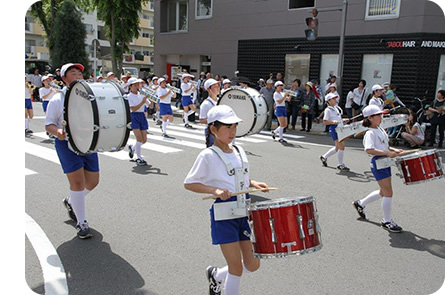 体操着に赤白帽子をかぶった子供達の鼓笛隊が歩行者天国になった道路をパレードしている写真