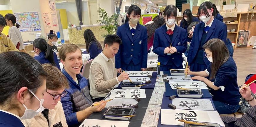 制服姿の女子生徒に教わりながら外国人の方々が書道を行っている様子の写真