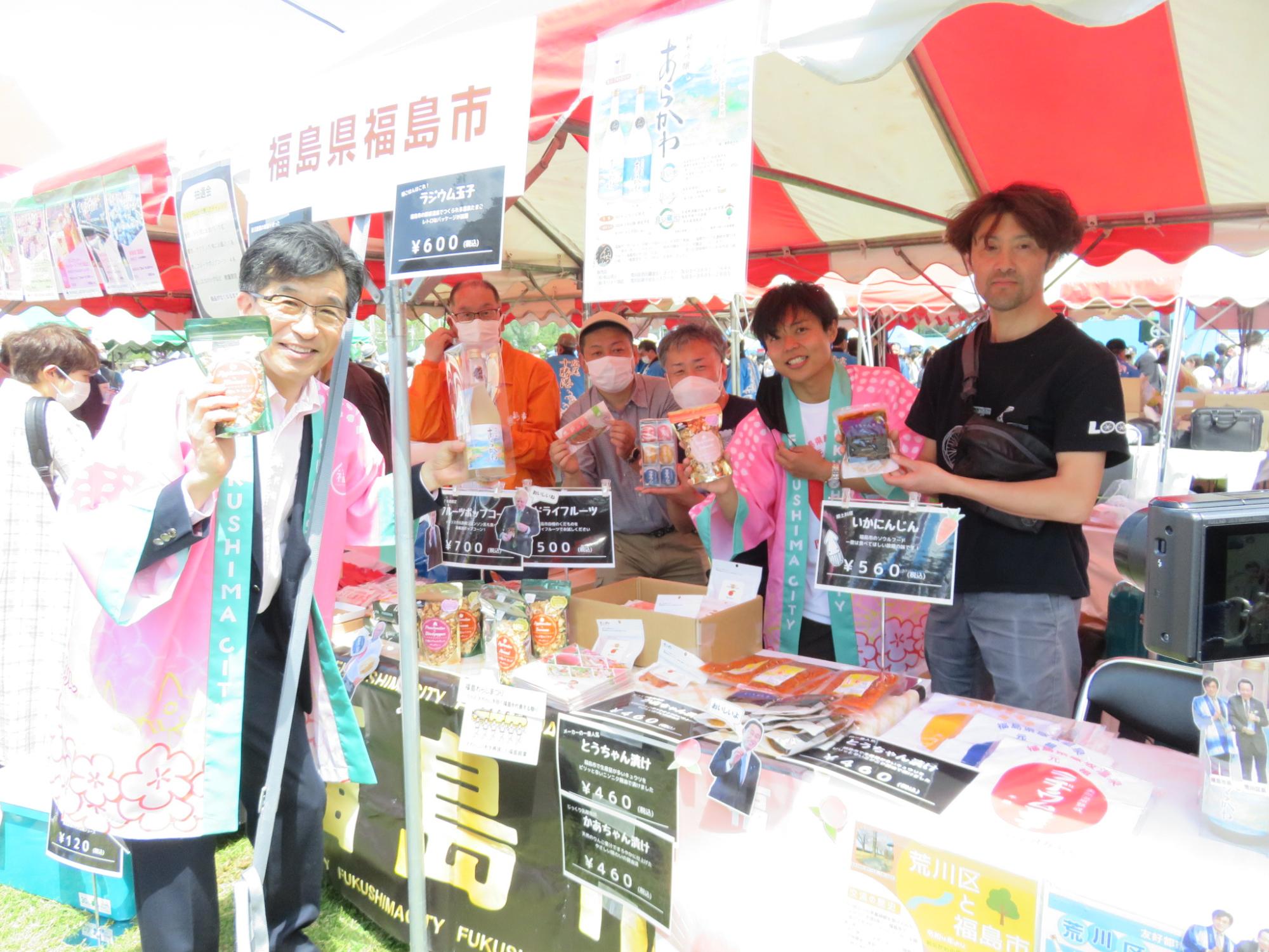 川の手荒川まつりでピンクの法被を着た市長や関係者達がそれぞれ特産品を持ちカメラに向け紹介している様子の写真