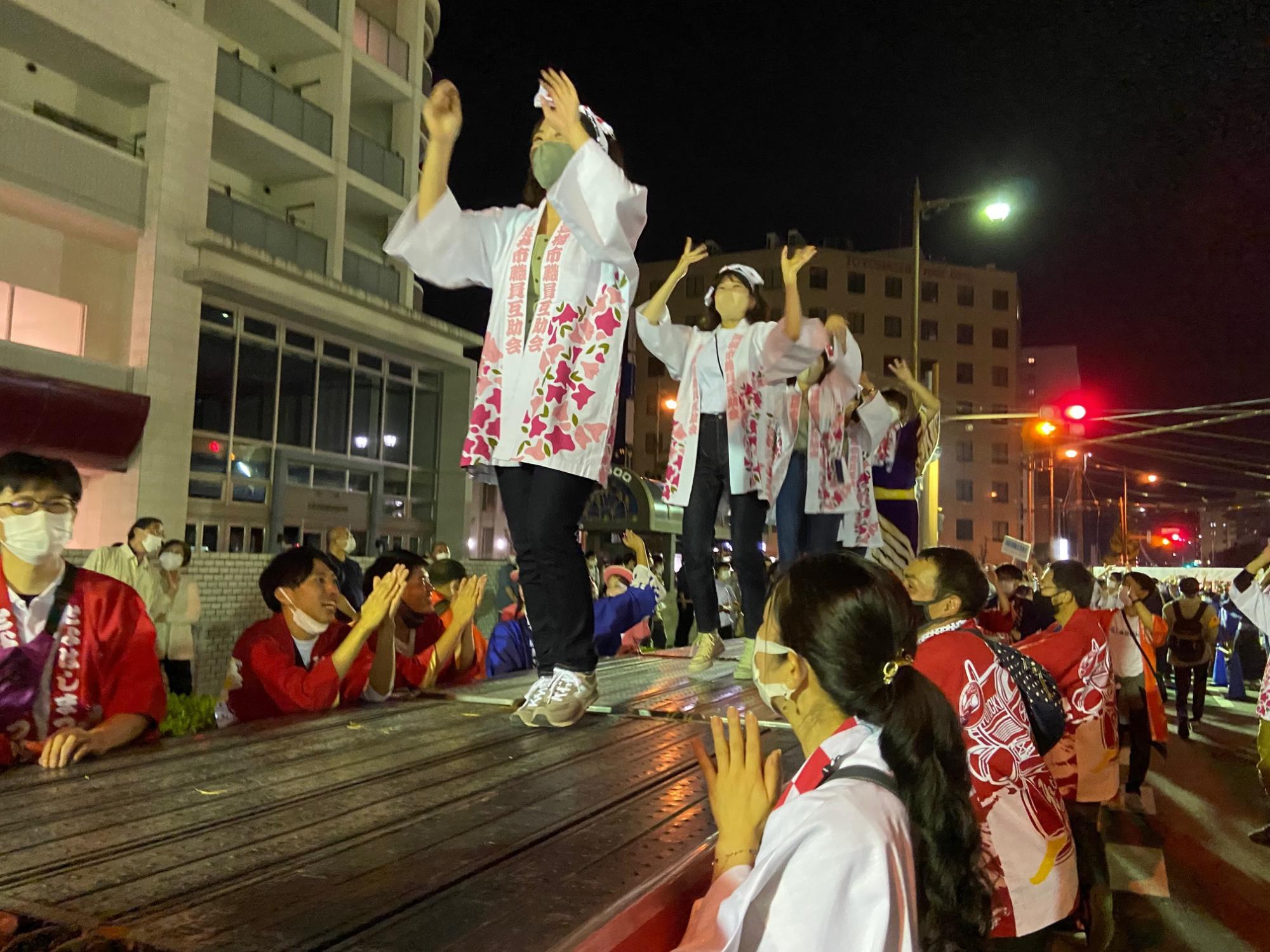 花柄の法被を着た女性達が舞台で踊っており、市役所職員が盛り上げている様子の写真