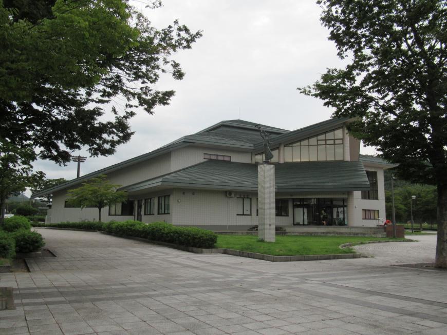 大きな木が立ち並んでいる奥に建つグレーの屋根の十六沼体育館の外観写真