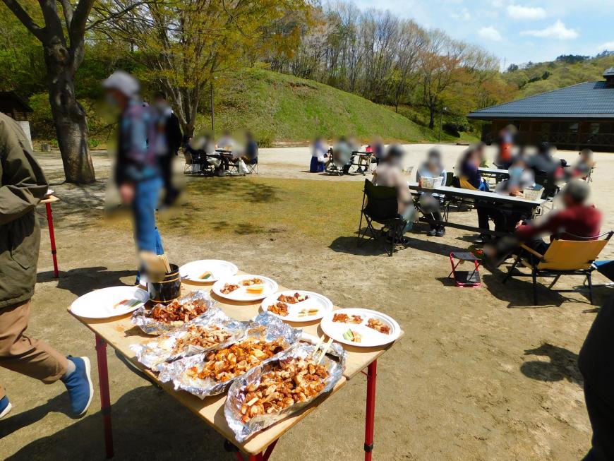 手前のテーブルに沢山の料理が並べられ、参加者達がグループに分かれて食事を楽しむ様子の写真