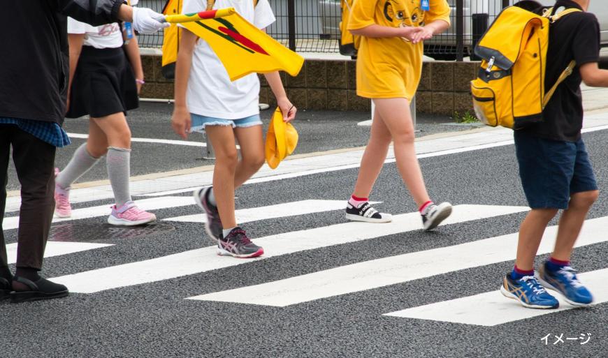 黄色い旗を持ったボランティアの方がいる横断歩道を渡っている子供たちの写真(イメージ)