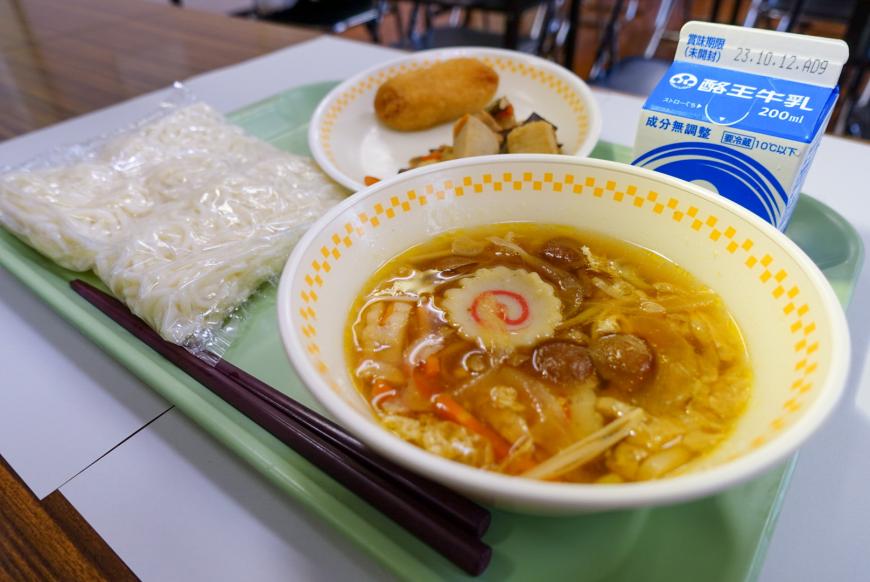 グリーンのトレーに袋に入った麺、お椀に入ったスープ、小皿に入ったおかず、牛乳パックが置かれている写真
