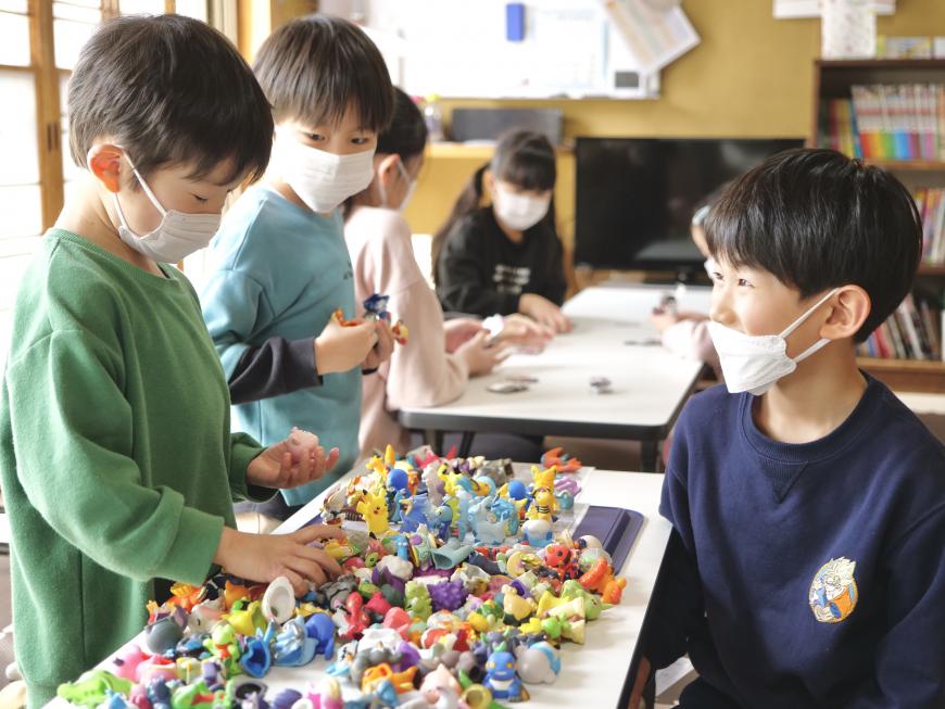 手のひらサイズの沢山の人形が置かれたテーブルから人形を選んでいる男の子と、その様子を見ている男の子の写真