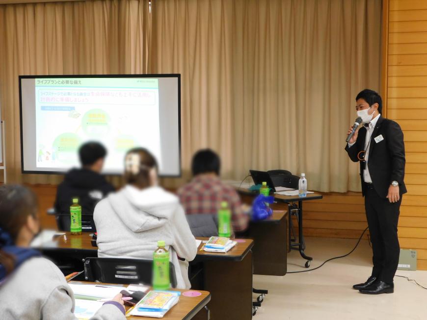 スクリーンに資料が写し出され、男性がマイクを持ち話をするのを参加者達が聞いている写真