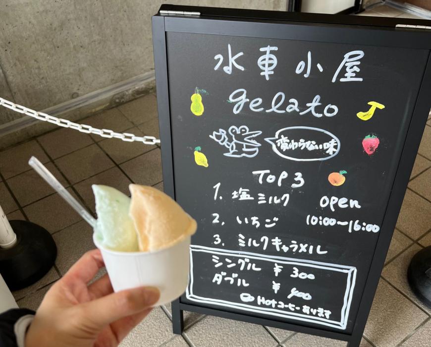 手に持ったカップに入ったジェラートと水車小屋のメニューがのった看板の写真