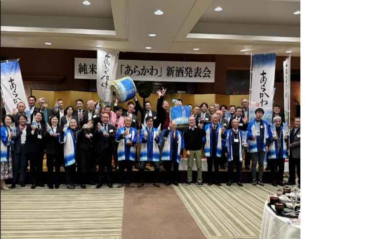 上に「純米吟醸「あらかわ」新酒発表会」と書かれ、「あらかわ」の旗と樽、関係者達の集合写真