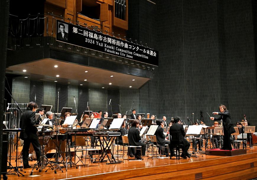 指揮者が指揮するステージ上でオーケストラ演奏が行われている写真
