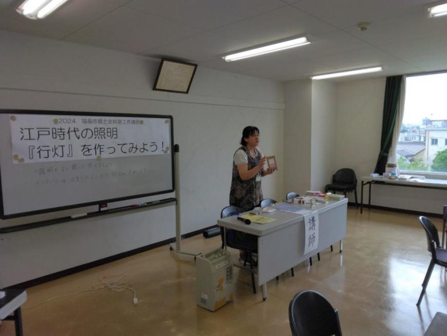 机に置かれた行灯を手に持ち受講者に作り方を教える講師を斜め前から撮影した写真