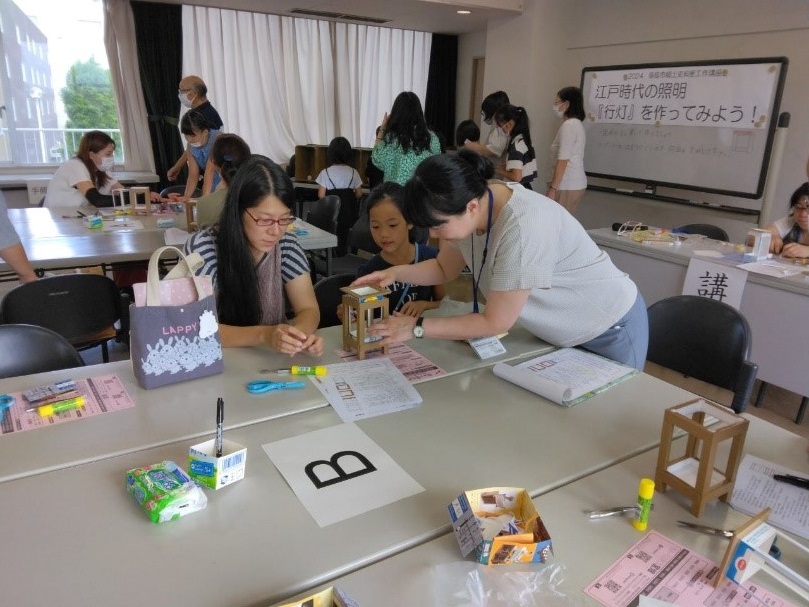 行灯の蓋の部分を慎重に作業している2人の女性と女の子を撮影した写真