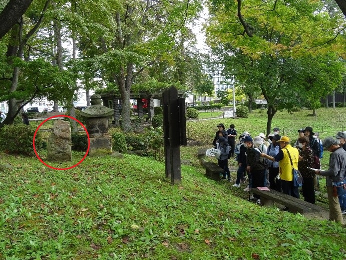 板碑の説明が書いてある看板の前で集まり講師の話を聞きながら観察する受講者たちを撮影している写真
