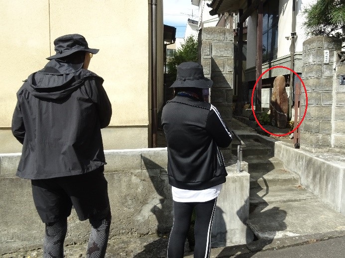 赤みがかった石碑をじっと見る黒い服の男女を後ろから撮影している写真