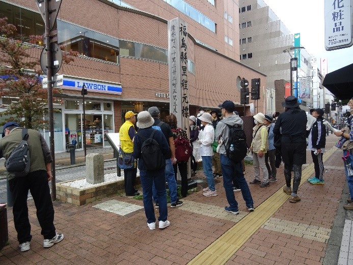街の真ん中に置かれた福島懸里程元標と彫られた大きな木の板の周りに集まる受講者たちを撮影した写真