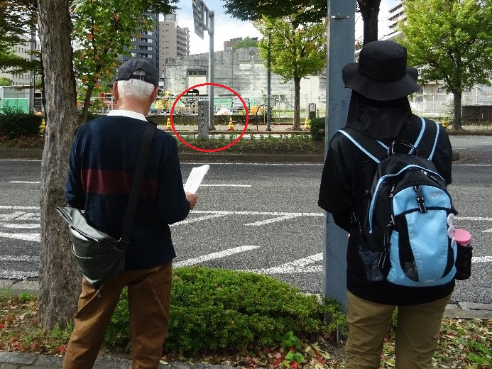 二車線道路の真ん中に置かれた福島城大手門跡の看板を道路脇から観察する男女の受講者を撮影した写真