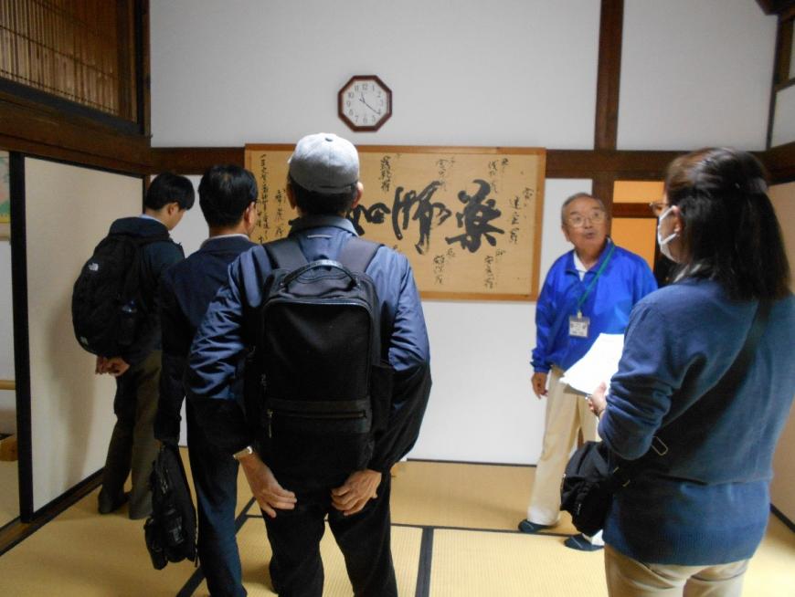 建物内に飾られた書き物について説明をするガイドと話を聞く受講者たちを撮影した写真