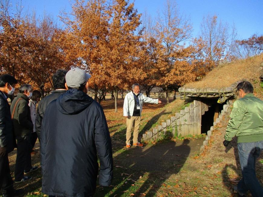 竪穴住居の入り口を手で指し示す講師と近づく参加者たちの写真
