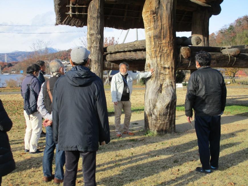 掘立柱建物の地面に刺さる太い木の柱に片手をつきながら話す講師と聞く参加者たちの写真