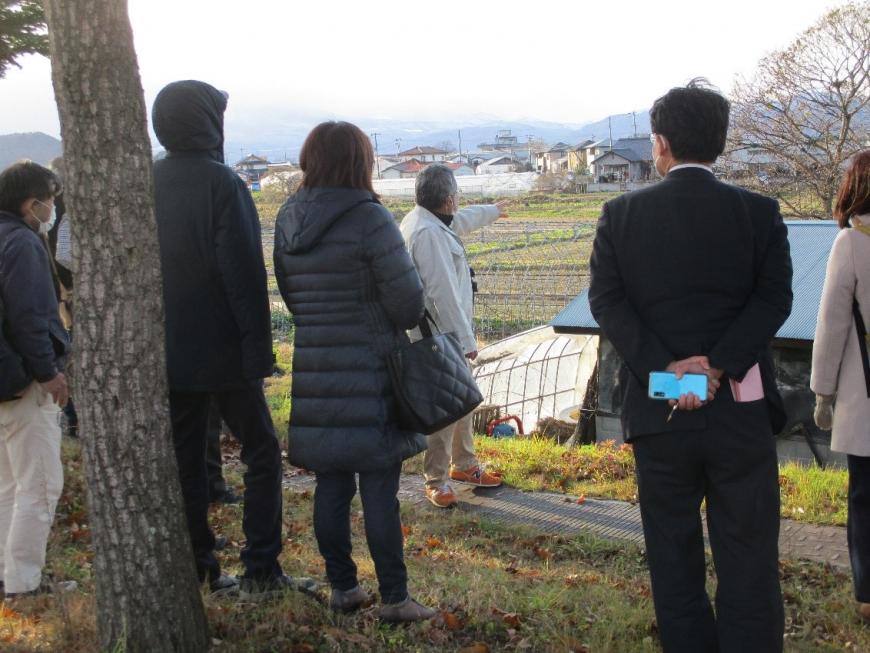 畑や民家が見渡せる広場から縄文時代に阿武隈川が流れていたであろう場所に指をさしながら話す講師と後ろで一緒に見る受講者たちの写真