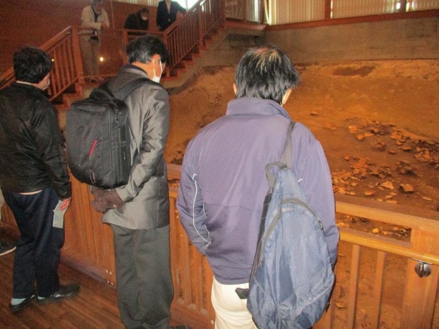 発掘調査跡の地面を柵越しにじっと見る2人の男性の後ろ姿を撮影している写真