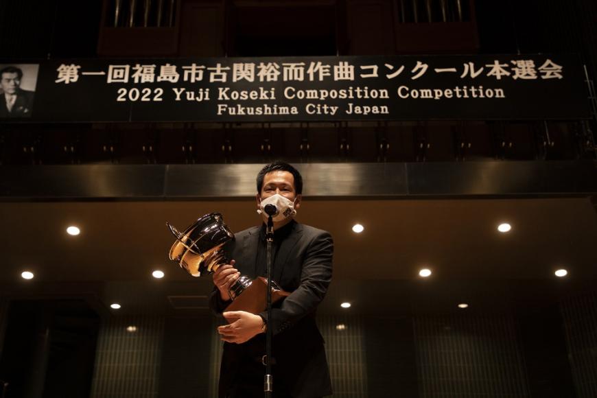 第一回古関裕而作曲コンクール本選会にて第一位を受賞した佐藤信人さんが大きなトロフィーを持ちマイクの前に立っている写真