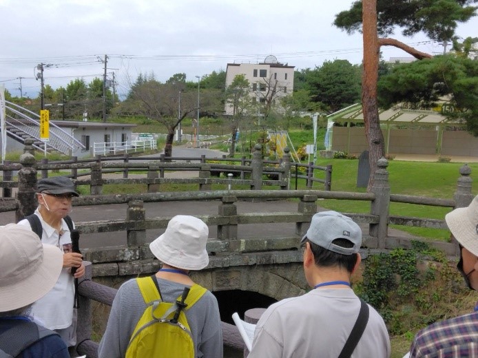 芝生が綺麗な噴水公園内に架けられた祓川の石橋を説明する講師と話を聞く受講者たちの写真