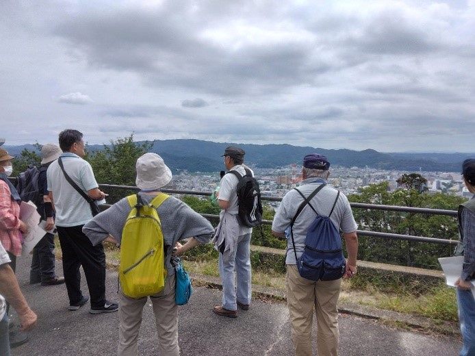 街並みを見渡せる第一展望台で福島盆地について説明をする講師と話を聞く受講者たちの写真